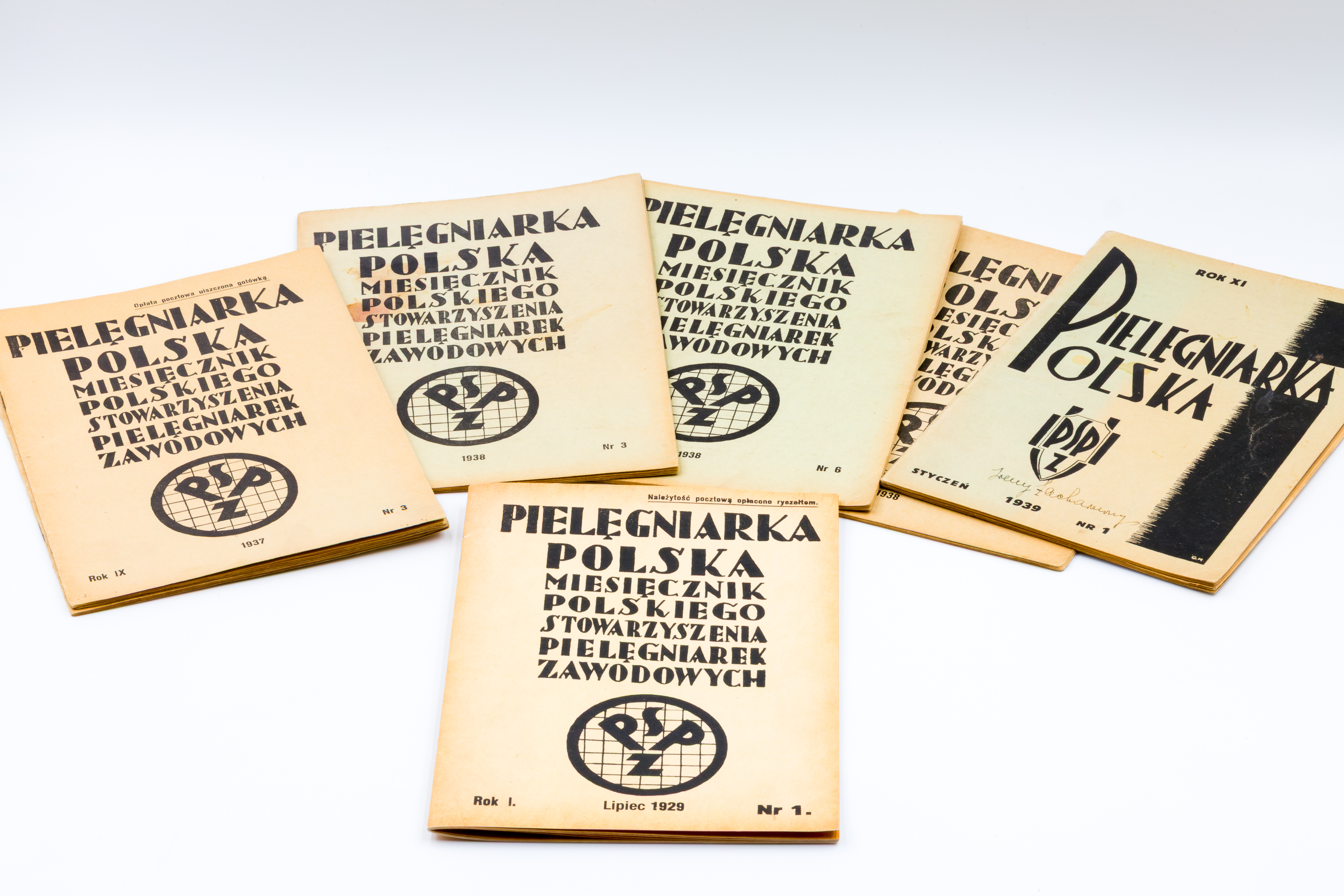 Na białym tle znajdują się ułożone w półokrąg stare, pożółkłe czasopisma. Są to różne numery  miesięcznika „Pielęgniarka Polska” wydawanego przez Polskie Stowarzyszenie Pielęgniarek Zawodowych.